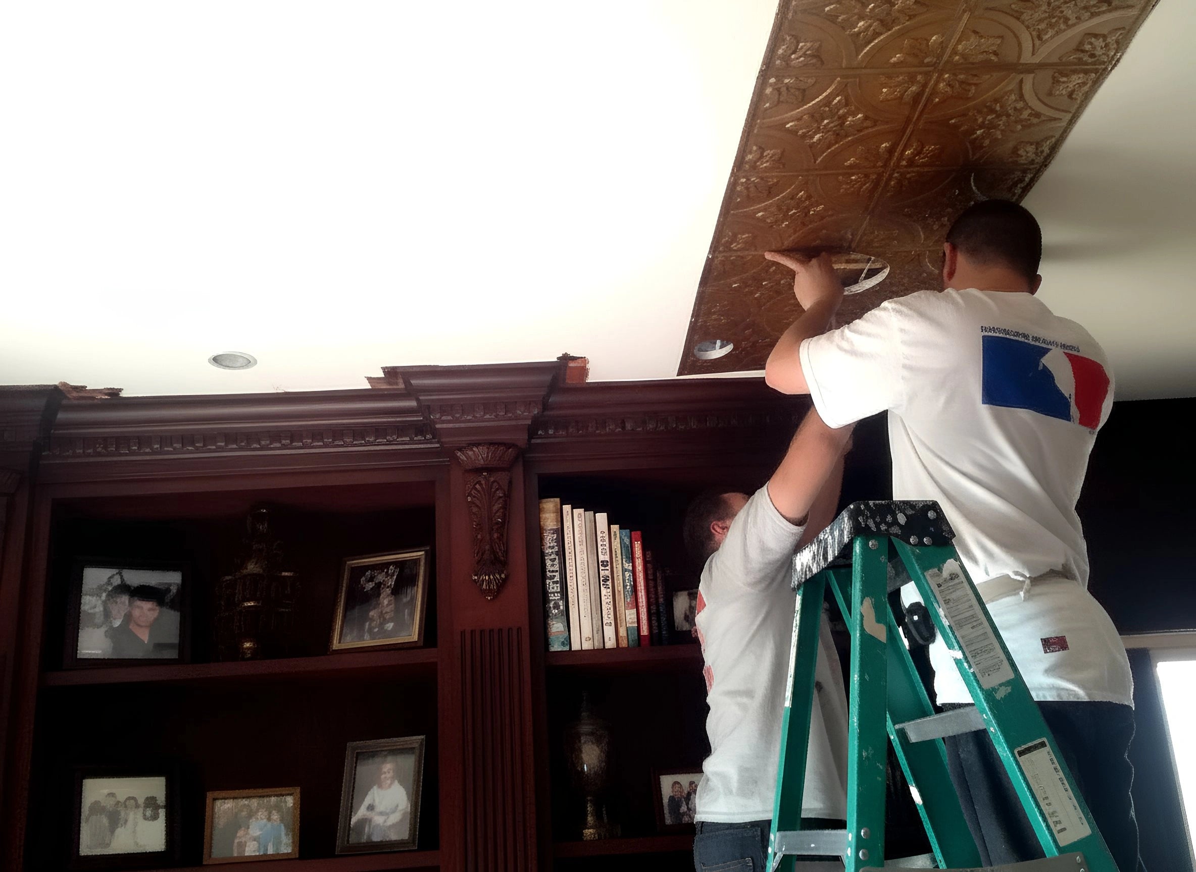 Person on a ladder working on a decorative ceiling in a room with bookshelves and framed pictures.